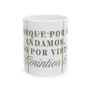Puede incluir: Una taza de cerámica blanca con un gráfico de arco de color dorado y texto en verde oscuro que dice: PORQUE POR FE ANDAMOS, NO POR VISTA 2 Corintios 5:7. El diseño está centrado en la superficie exterior del recipiente cilíndrico.