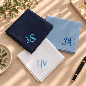 May include: Three folded square handkerchiefs in navy blue, light blue, and white. Each features embroidered monogram initials in light blue thread: VBS on the navy, JA on the light blue, and L/V on the white. The fabric has a visible weave.