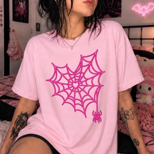 Pode incluir: Uma t-shirt de manga curta rosa claro com um grande gráfico rosa choque de uma teia de aranha em forma de coração. Uma pequena aranha está pendurada na parte inferior do design. O gráfico tem uma aparência brilhante e texturizada.