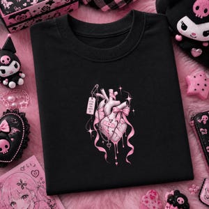 May include: A black t-shirt featuring a central graphic of an anatomical heart in shades of pink and white. The heart is adorned with a pink bow, a bandage, a safety pin, and a small tag with the text Still Feel Too Much and a heart symbol.