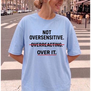 May include: A light blue short-sleeve t-shirt featuring black text that reads NOT OVERSENSITIVE. OVERREACTING. OVER IT. The word OVERREACTING has a red line struck through it, and the word OVER IT. has a red line underlined beneath it.
