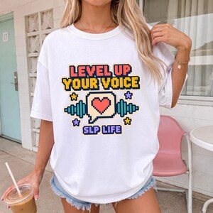 May include: A white short-sleeve t-shirt featuring a pixelated graphic with the text LEVEL UP YOUR VOICE and SLP LIFE. The design includes a central speech bubble with a red heart, flanked by teal sound waves and small purple and yellow stars.
