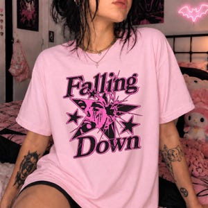 Puede incluir: Una camiseta de manga corta de color rosa claro con un estampado gráfico en negro y rosa oscuro. El diseño incluye una estrella grande, un rostro estilizado y el texto "Falling Down" en una fuente decorativa y negrita en el pecho.