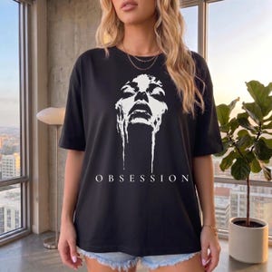 May include: A black short-sleeve t-shirt featuring a white graphic print of a face with dripping details and the word OBSESSION printed in white capital letters below the graphic. The shirt has a crew neckline and a relaxed, loose-fitting silhouette.