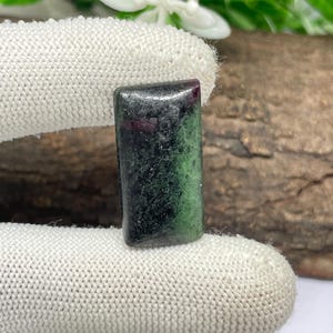 Op de afbeelding: Een rechthoekige cabochon edelsteen met een mix van diep bosgroene en zwarte zoisiet mineraalmatrix met kleine, ingebedde vlekken van levendig magentarode robijn, gepolijst tot een glad, reflecterend oppervlak met afgeronde randen.