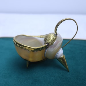 Puede incluir: Un cuenco decorativo hecho de una concha natural con acabado iridiscente. La concha está montada sobre tres patas metálicas doradas en punta y presenta un borde dorado festoneado, un adorno floral de metal y un asa metálica curva.