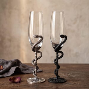 May include: Two tall champagne flutes with clear glass bowls. Each stem features a detailed snake coiled around a textured branch-like base. One flute has a metallic silver finish, while the other has a matte black finish with a small red eye detail.