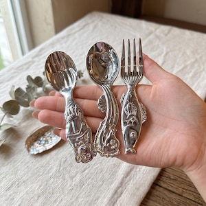May include: A set of three silver-toned metal utensils consisting of two spoons and one fork. Each handle features a detailed embossed design of sea creatures, including a monkey, a seahorse, and a fish, with a polished reflective finish.