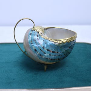 Puede incluir: Un cuenco de concha de nautilo con un exterior pulido iridiscente en azul y blanco, un borde de metal dorado, un adorno de filigrana en la parte superior, un asa curva y tres pequeñas patas de metal que sostienen la base.