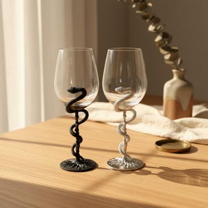 May include: Two clear glass wine glasses with stems shaped like coiled snakes. One glass features a matte black snake stem, while the other has a metallic silver snake stem. Both stems extend from a circular base to the bowl of the glass.
