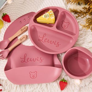 May include: A dusty rose silicone feeding set featuring a three-section plate, a bowl, a bib, a spoon, and a fork. The plate, bowl, and bib are embossed with the name Lewis and a bear face icon. The utensils have wooden handles and silicone tips.