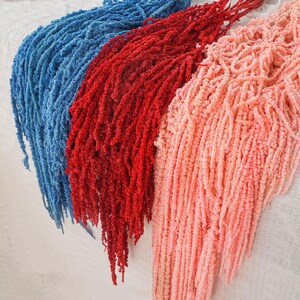 May include: Three bundles of dried amaranthus hanging downwards, featuring long, cascading, textured flower strands in vibrant shades of blue, deep red, and soft peach. Each bundle consists of numerous thin, delicate, and granular stems.