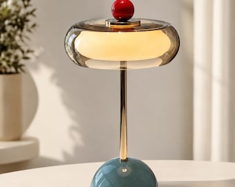 Könnte beinhalten: Eine Tischleuchte mit einem kuppelförmigen, blaugrünen Sockel, einem schlanken goldfarbenen Metallstiel und einem breiten, durchscheinenden grauen Glasschirm, der einen weißen Lichtdiffusor umschließt. Eine kleine, glänzende rote Kugel sitzt oben auf dem Glasschirm.