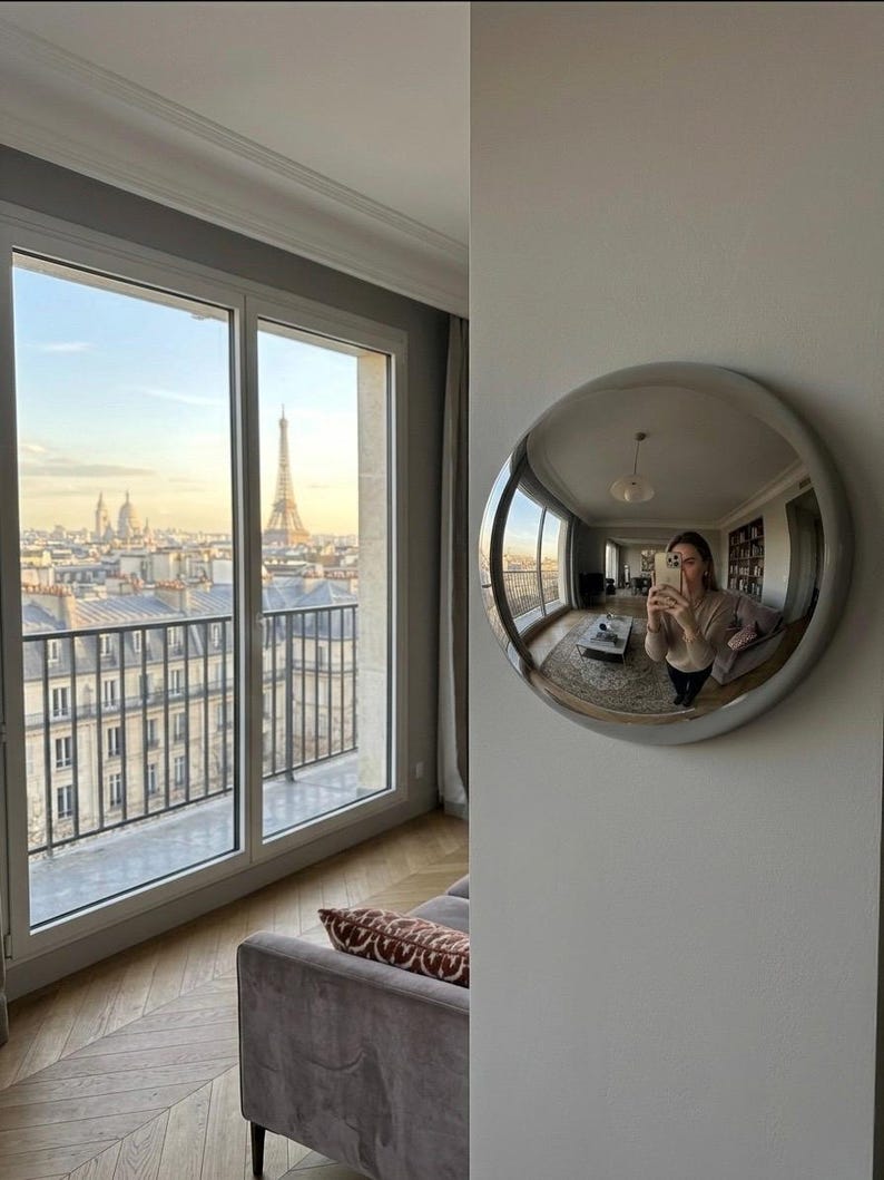 Peut inclure: Un miroir mural convexe circulaire doté d'un cadre argenté lisse, fixé sur un mur blanc. Le miroir reflète une vue grand-angle d'un espace de vie intérieur, incluant des meubles et une personne tenant un smartphone pour capturer le reflet.