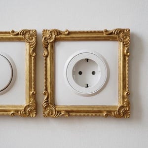 Op de afbeelding: Een wit stopcontact en een lichtschakelaar, elk omlijst door een vierkante, goudkleurige rand met decoratieve hoekdetails. De lijsten zijn direct op een vlakke, effen witte muur gemonteerd en omsluiten de elektrische armaturen.