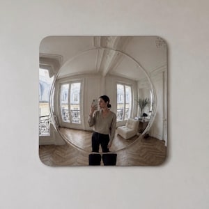 May include: A square wall mirror with rounded corners featuring a large, circular convex centre that creates a wide-angle, distorted reflection of a room with light-coloured walls, wooden floors, two windows, and a piece of furniture.