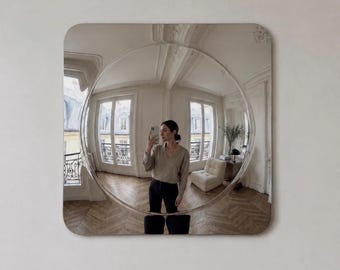 May include: A square wall mirror with rounded corners featuring a large, circular convex centre that creates a wide-angle, distorted reflection of a room with light-coloured walls, wooden floors, two windows, and a piece of furniture.