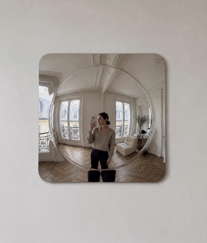 May include: A square wall mirror with rounded corners featuring a large, circular convex center that creates a wide-angle, distorted reflection of a room with light-colored walls, wooden floors, two windows, and a piece of furniture.
