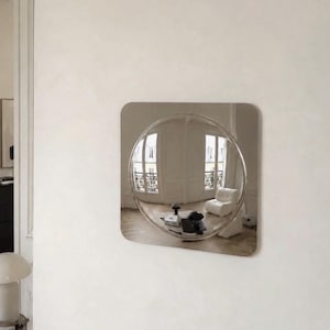 May include: A square wall mirror with a polished metallic finish featuring a large, circular, convex center section that creates a wide-angle, distorted reflection of a room with white walls, a white armchair, and a small table.