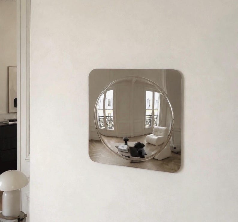 May include: A square wall mirror with a polished metallic finish featuring a large, circular, convex center section that creates a wide-angle, distorted reflection of a room with white walls, a white armchair, and a small table.