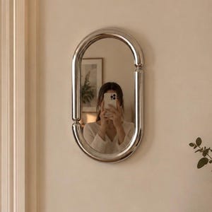 Peut inclure: Un miroir mural ovale doté d'un cadre épais en chrome poli au design tubulaire distinct. Le cadre se compose de deux sections latérales incurvées reliées par des joints, créant une bordure métallique arrondie et continue autour du verre.