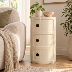 May include: A cylindrical cream-coloured storage unit featuring three stacked drawers, each equipped with a circular cutout handle. The unit has a smooth, glossy finish and a rounded profile, providing a compact vertical storage solution for home.