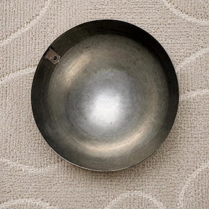 K&ouml;nnte beinhalten: Eine kreisf&ouml;rmige, tiefe Metallschale mit einer dunklen, verwitterten Patina und einer glatten, reflektierenden Innenfl&auml;che. Eine kleine, rechteckige Metalllasche mit einem einzelnen kreisf&ouml;rmigen Loch ist am inneren oberen Rand der Schale befestigt.