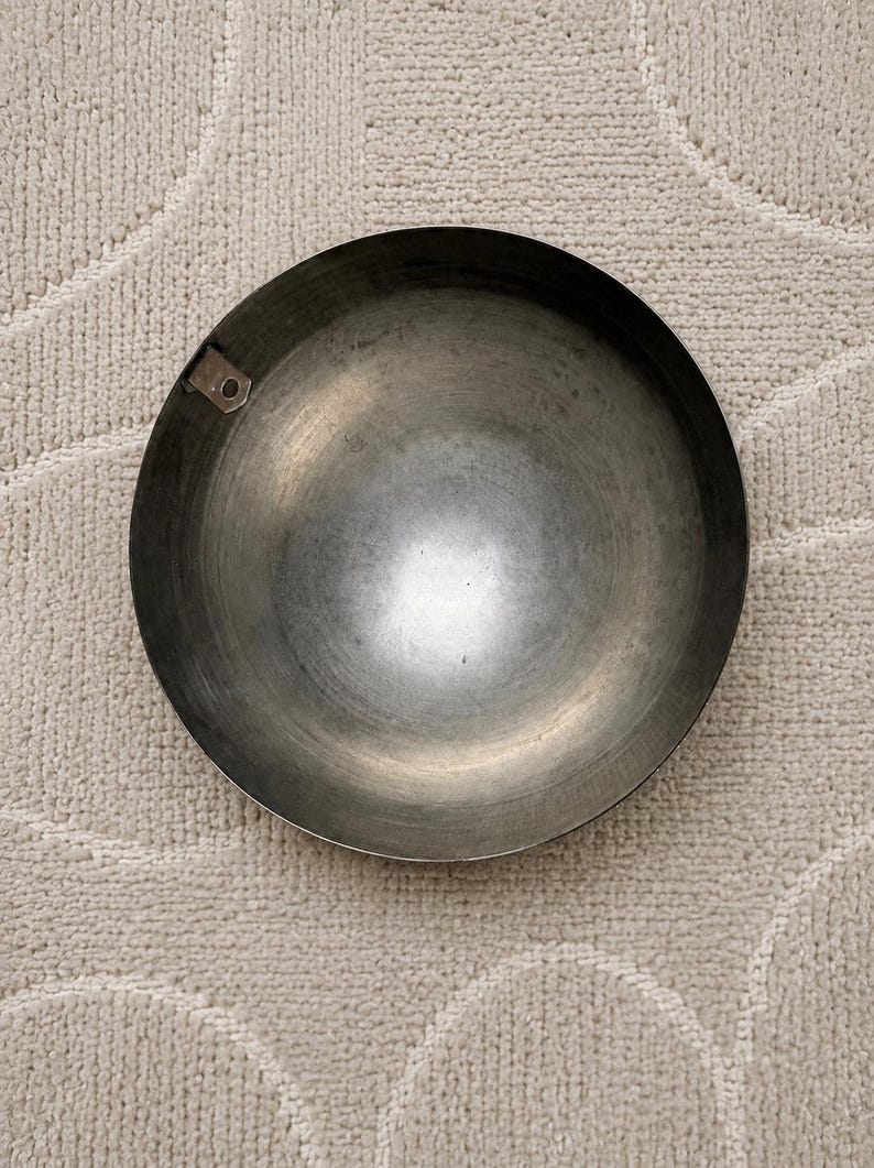 K&ouml;nnte beinhalten: Eine kreisf&ouml;rmige, tiefe Metallschale mit einer dunklen, verwitterten Patina und einer glatten, reflektierenden Innenfl&auml;che. Eine kleine, rechteckige Metalllasche mit einem einzelnen kreisf&ouml;rmigen Loch ist am inneren oberen Rand der Schale befestigt.