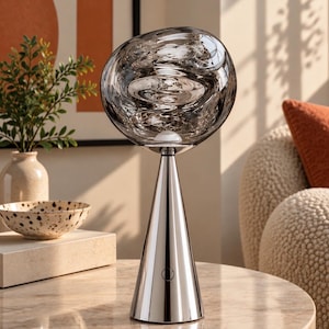 Può includere: Una lampada da tavolo con base conica in argento lucido e paralume sferico in vetro fumé con una texture vorticosa e ondulata. La lampada include un piccolo pulsante di accensione circolare sulla parte anteriore della base metallica.