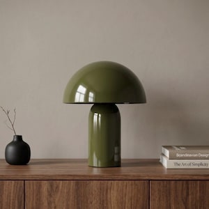 Può includere: Una lampada da tavolo verde oliva lucido con paralume a cupola a forma di fungo e base cilindrica. La lampada è posizionata tra un piccolo vaso nero contenente un rametto sottile e una pila di due libri intitolati Scandinavian Design e The Art of Simplicity.