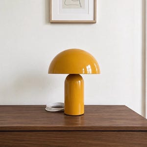 May include: A mustard yellow table lamp with a smooth, glossy finish. The lamp features a wide, dome-shaped shade resting on a cylindrical base. A white power cord is coiled at the base of the lamp, which has a simple, rounded silhouette.