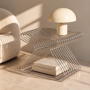 May include: A chrome-finished metal side table with a geometric, wire-frame structure featuring a central inward curve. A cream-coloured mushroom-shaped table lamp sits on the top surface, and two cream-coloured books are stacked on the bottom shelf.