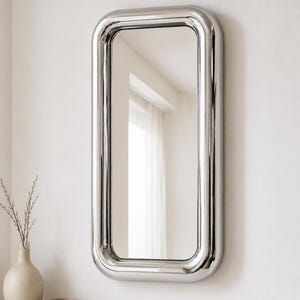 Può includere: Uno specchio da parete rettangolare con una spessa cornice in cromo lucido dagli angoli arrotondati. La superficie riflettente è chiara e liscia, racchiusa nel bordo metallico che crea un profilo arrotondato distintivo sulla parete.