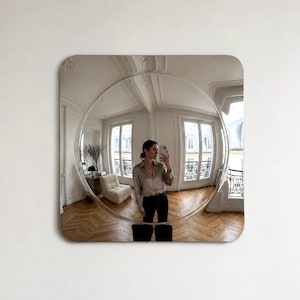 Peut inclure: Un miroir mural carré aux coins arrondis doté d'un grand centre convexe circulaire qui crée une réflexion grand-angle déformée d'une pièce avec parquet, murs blancs, fenêtres et une personne tenant un smartphone.