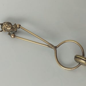 May include: A brass-toned metal tool featuring a sculpted human head at one end, two long thin arms connected by a central pivot, and a circular ring at the opposite end attached to a small, flat, round disc component with a smooth finish.
