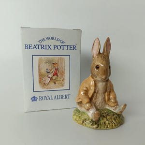 Peut inclure: Une figurine en céramique représentant un lapin brun portant une veste beige, assis sur une base verte, accompagnée d'une boîte en carton blanche marquée The World of Beatrix Potter et Royal Albert avec une illustration d'un canard et d'un renard.