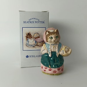 Op de afbeelding: Een keramieken kattenbeeldje met een roze mutsje, een blauwe sjaal en een groen schort over een roze rok met rode stippen, terwijl het een mandje vasthoudt. Daarnaast staat een witte kartonnen doos met de tekst The World of Beatrix Potter en Royal Albert.