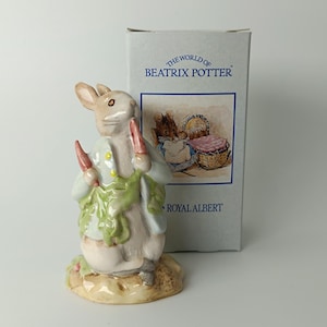 Op de afbeelding: Een porseleinen beeldje van Pieter Konijn in een lichtblauw jasje met een wortel, staand op een voetstuk. Daarachter staat een grijze doos met het opschrift The World of Beatrix Potter en Royal Albert, voorzien van een illustratie.
