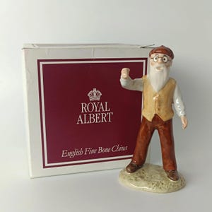 Op de afbeelding: Een Royal Albert English Fine Bone China porseleinen beeldje van een ouder persoon met een witte baard, gekleed in een bruine pet, geel vest en bruine broek, staand op een textuurrijke voet naast een witte doos met bordeauxrode opdruk.