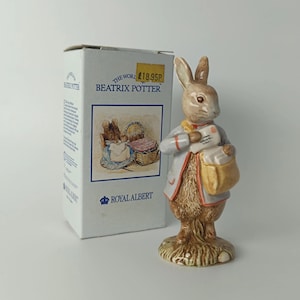 Peut inclure: Une figurine en céramique représentant un lapin brun vêtu d'une veste grise, portant une sacoche beige et une enveloppe blanche. À côté se trouve une boîte en carton blanche marquée The World of Beatrix Potter et Royal Albert.