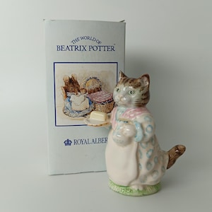 May include: A porcelain figurine of a cat wearing a patterned dress and apron, holding a small plate with a piece of butter. Beside it stands a white cardboard box labelled The World of Beatrix Potter and Royal Albert with an illustration.
