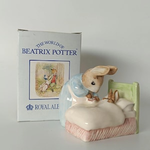 Peut inclure: Une figurine en porcelaine représentant un lapin en veste bleue bordant un plus petit lapin dans un lit à tête de lit vert pâle et literie blanche, accompagnée d'une boîte en carton blanche marquée The World of Beatrix Potter et Royal Albert.