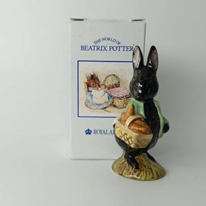 Op de afbeelding: Een keramieken beeldje van een zwart konijn in een lichtgroen jasje met een rieten mand vol brood, staand op een getextureerde voet. Daarachter staat een witte doos met de tekst THE WORLD OF BEATRIX POTTER en ROYAL ALBERT.