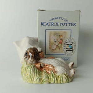 Könnte beinhalten: Eine Keramikfigur eines braun-weißen Kaninchens, das im grünen Gras liegt und eine weiße Schlafmütze trägt. Hinter der Figur steht ein weißer Karton mit dem Text THE WORLD OF BEATRIX POTTER und einem runden 100-Jahre-Jubiläumsemblem.