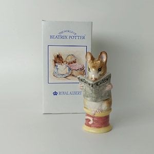 Op de afbeelding: Een keramieken beeldje van een bruine muis die een krant leest met de titel The Tailor & Cutter, zittend op een roze en gele garenklos. Daarachter staat een witte rechthoekige doos met het opschrift The World of Beatrix Potter en Royal Albert.