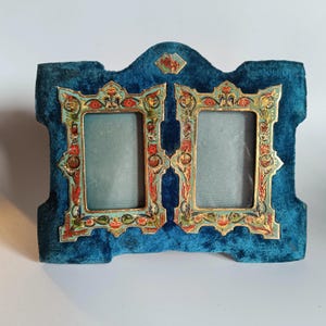 May include: A double photo frame featuring a deep blue velvet backing and two rectangular openings. The frame is decorated with ornate, multi-coloured floral patterns in shades of red, green, and gold, with a small diamond-shaped accent at the top.