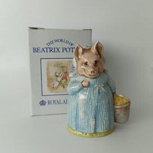 Peut inclure: Une figurine en céramique représentant un cochon vêtu d'une robe à motifs bleus et blancs et d'un bonnet, tenant un petit seau jaune. Derrière la figurine se trouve une boîte en carton blanche marquée The World of Beatrix Potter.
