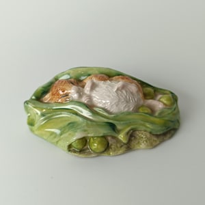 Peut inclure: Une figurine en céramique représentant un petit chaton orange et blanc dormant à l'intérieur d'une cosse de petit pois verte. La cosse présente des feuilles vertes texturées et plusieurs petits pois ronds nichés autour du chaton, le tout verni.