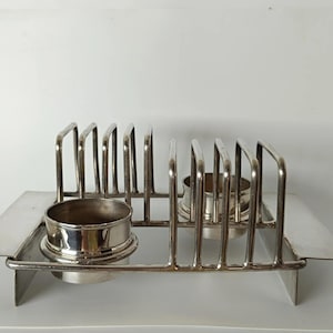 May include: A silver-toned metal toast rack featuring a rectangular base with flat side panels and a series of vertical wire dividers. Two circular, polished metal containers are integrated into the structure, positioned within the rack frame.
