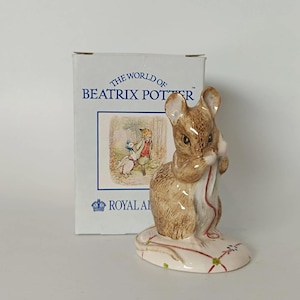 May include: A porcelain figurine of a brown mouse holding a red thread, standing on a base with a red and green pattern. Behind it is a white box labelled THE WORLD OF BEATRIX POTTER with an illustration of a fox and a duck in a forest.